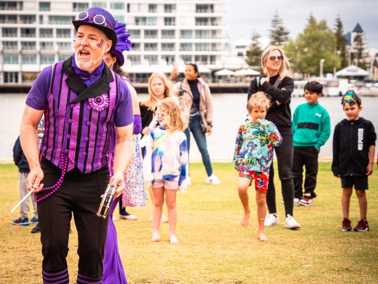 Image of A man in a purple costume and hat entertains children and adults outdoors near a waterfront. The group stands on grass with modern buildings, trees, and cloudy skies in the background.