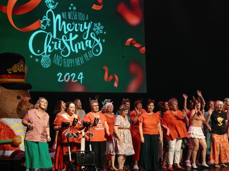 Image of A group of people in festive outfits sing on stage in front of a screen that reads We wish you a Merry Christmas and Happy New Year 2024. A large snowman decoration is visible on the left.