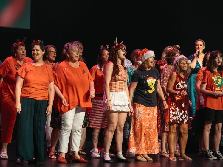 Image of A group of women dressed in festive, colourful clothing sing together on stage. Some wear reindeer antlers, Santa hats, or Christmas-themed outfits, creating a lively holiday atmosphere.