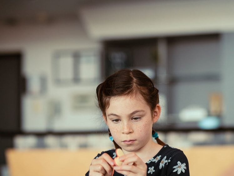 Image of A young girl in a black floral top focuses on crafting at a table. Colourful heart-shaped decorations with letters hang above her in the background.