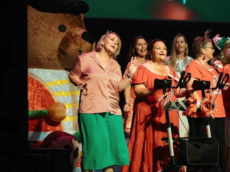 Image of A group of women sing and perform on stage, some wearing red and white outfits, standing beside a festive bear decoration and a walking frame decorated with ribbons. The background is dark with stage lighting.