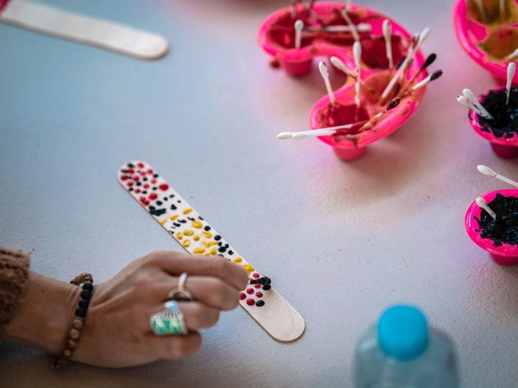 Image of A hand decorates a wooden craft stick with colourful paint dots using a cotton bud. Nearby are pink paint trays holding buds and various paint colours.
