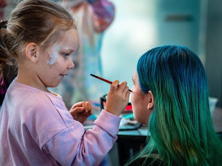 Image of A young girl with face paint carefully applies make-up to an adult with long blue and green hair. The girl is holding a brush and focussing on painting the adult's face.