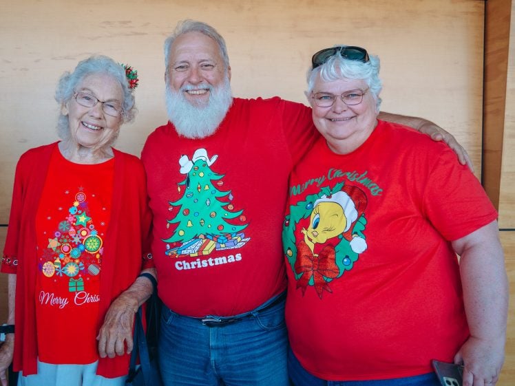 Image of Three smiling older adults stand together wearing festive red Christmas shirts, posing indoors against a wooden wall. The central person has a white beard, and everyone looks cheerful and happy.