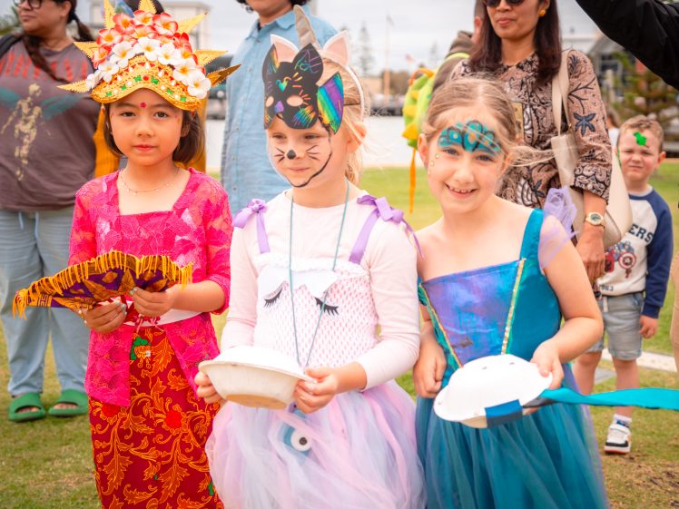 Image of Three young girls in colourful costumes stand outdoors at a festival. One wears a traditional red and gold outfit, another is dressed as a cat with a face mask and tutu, and the third wears a blue dress and face paint. Adults and children are in the background.