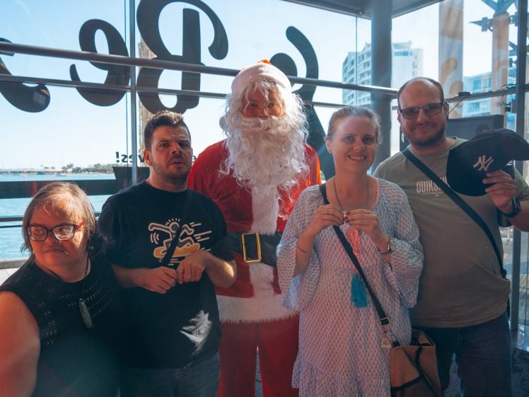 Image of Five adults, including a person dressed as Father Christmas, stand together smiling inside a glass building with sunlight streaming in. A waterfront and buildings are visible outside through large windows.