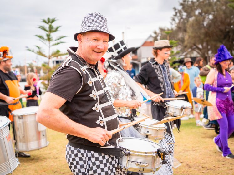 Image of A smiling man in a chequered hat plays the snare drum with a festive group of drummers and people in colourful costumes parading outdoors on a grassy area, with a crowd of onlookers in the background.