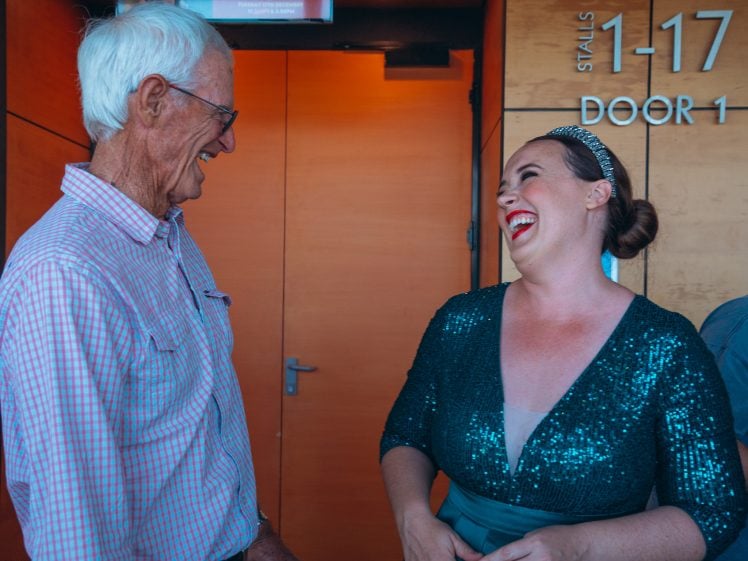 Image of An older man and a woman in a sparkly green dress are laughing together in a corridor, standing near a wooden door labelled Stalls 1-17 Door 1.