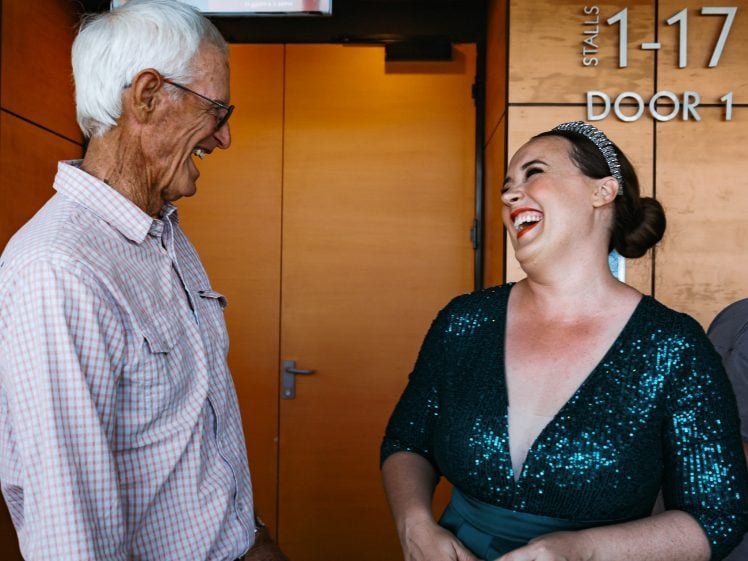 Image of An older man and a woman in a sparkly green dress laugh together in front of a wooden door, with signage reading STALLS 1–17 and DOOR 1 visible on the wall behind them.