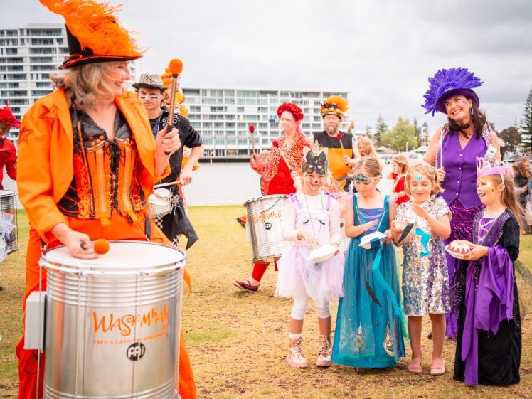 Image of A group of people in colourful costumes play drums and celebrate outdoors. Children in fancy dress stand in front, smiling, whilst adults in vibrant outfits and hats play instruments. Modern buildings and cloudy sky are in the background.