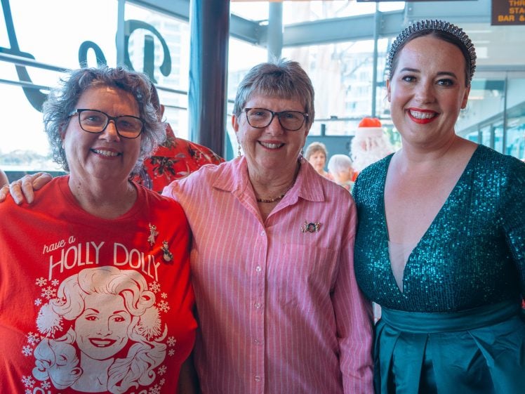 Image of Three women smiling and posing together indoors. The woman on the left wears a red Dolly Parton holiday jumper, the middle woman has short grey hair and glasses with a pink striped shirt, and the woman on the right wears a green sparkly dress.