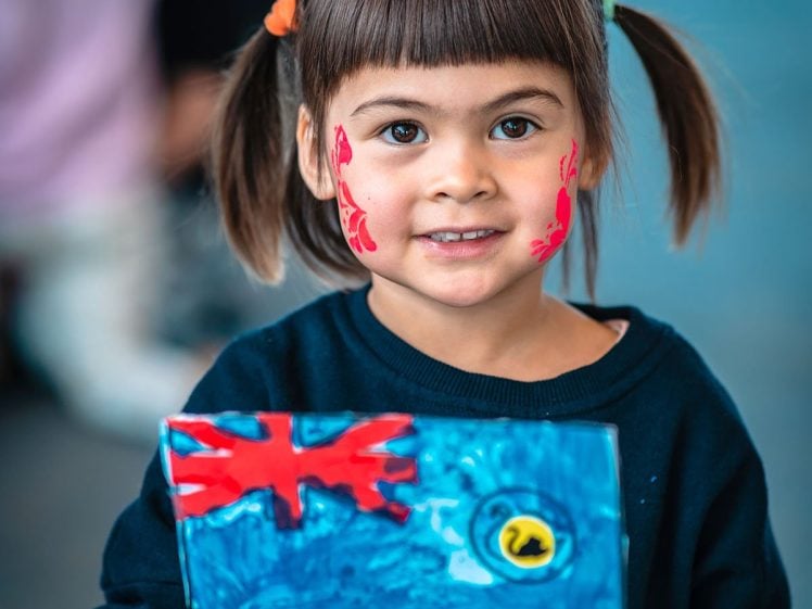 Image of A young child with plaits and red face paint on her cheeks smiles whilst holding a handmade blue flag. She wears a dark shirt with a colourful owl design. The background is blurred, showing other children.