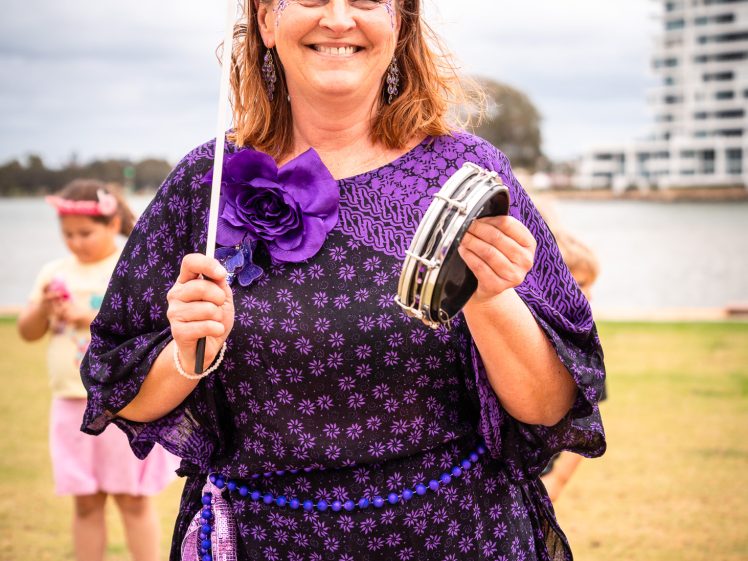 Image of A smiling woman in a purple outfit and floral headpiece holds a tambourine and a stick outdoors. Children and a waterfront with modern buildings are in the background.