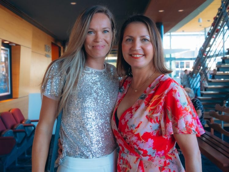 Image of Two women smiling and standing together indoors. One wears a shiny silver sequin top and white skirt; the other wears a red floral dress. They are in a well-lit area with seats and a staircase in the background.