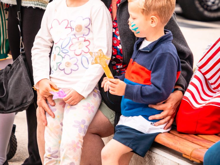 Image of An older woman with grey hair and sunglasses smiles whilst sitting with two young children, both with painted faces, on a bench outdoors. The girl wears floral clothes; the boy has a striped shirt and holds a yellow toy.
