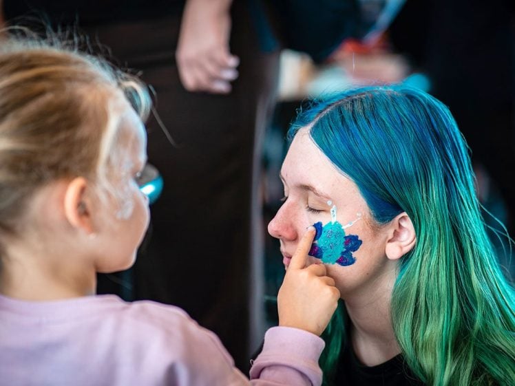 Image of A young child paints a colourful design on the face of a person with long blue and green hair, who has their eyes closed and sits still, enjoying the face painting.