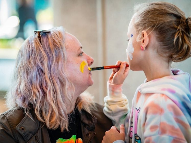 Image of A young girl paints a yellow crescent on a woman’s cheek while both have face paint and smile. The woman has fair hair and sunglasses on her head; the girl wears a pastel hoodie and holds a paintbrush close to the woman.