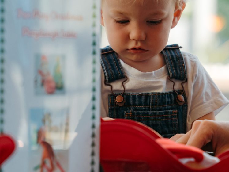 Image of A young child with blond hair wearing denim dungarees and a white shirt focuses on playing at a table with toy items and small containers. A sign with pictures and text is visible in the foreground.