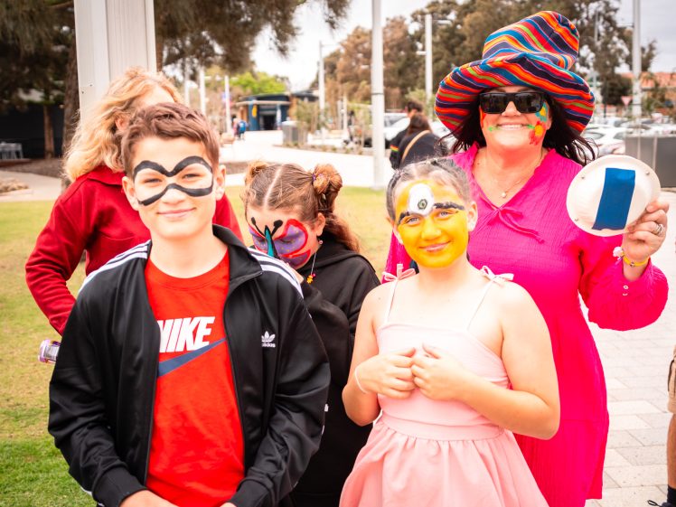 Image of A group of children and an adult pose outdoors, all with colourful face paint. The adult wears a bright, multicoloured hat and pink dress. The kids stand in front, smiling, holding plates, with trees and a park path in the background.