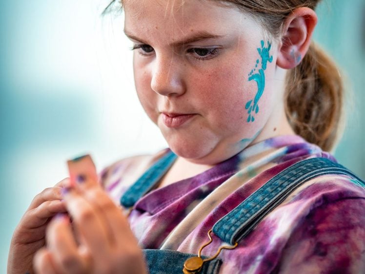 Image of A young girl with a blue lizard painted on her cheek looks intently at an object in her hand. She wears a tie-dye shirt and denim dungarees, and has a small blue shape painted on her wrist.