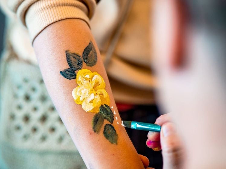 Image of A person paints a yellow flower with green leaves on someone's forearm using a small brush. The artist wears a beige jumper with a patterned sleeve. The focus is on the painted tattoo and the hand holding the brush.