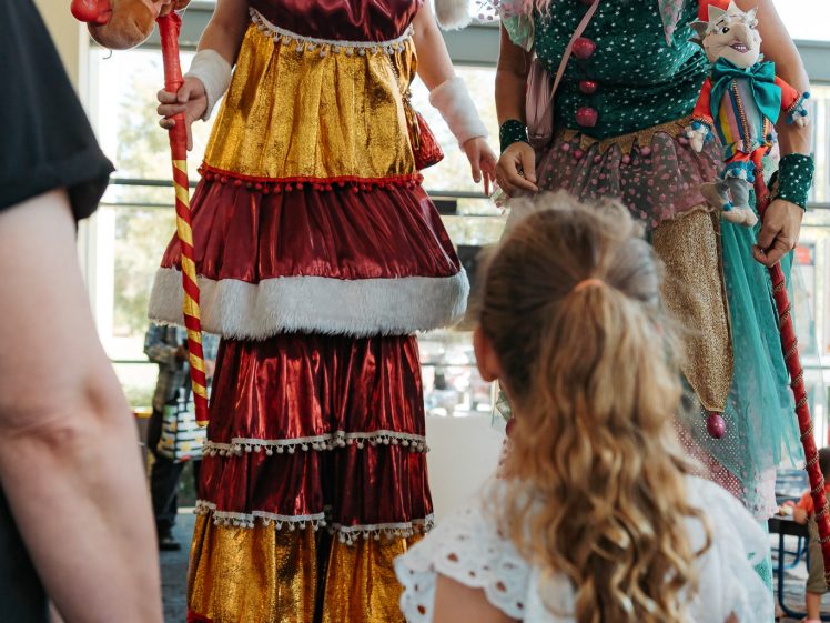 Image of Two women in colourful, festive costumes on stilts interact with a young girl holding hands with an adult. The scene takes place indoors with large windows in the background.
