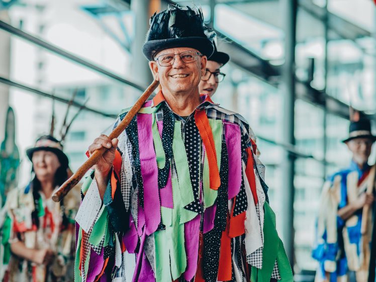 Image of An older man in a colourful costume with ribbons and a feathered hat smiles and holds a stick. He stands indoors with others in similar outfits blurred in the background.