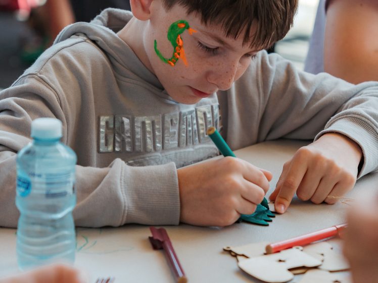 Image of A boy with a green face painting of a dinosaur concentrates on colouring a paper cut-out at a table, surrounded by coloured markers and a water bottle. Other children are visible nearby.