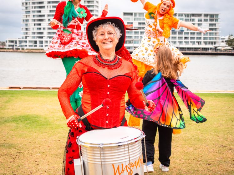 Image of A smiling woman in a red costume plays a drum labelled Wasamba on grass, while two performers on stilts in colourful fruit costumes and a child in butterfly wings are in the background near a waterfront and modern buildings.