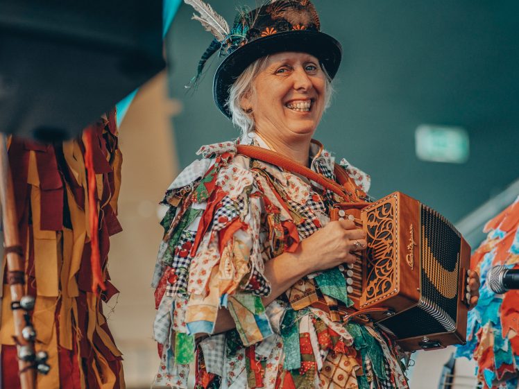 Image of A smiling person wearing a colourful patchwork costume and a decorated hat with feathers plays an accordion on stage.