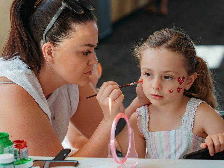 Image of A woman carefully paints a young girl's face with a brush, adding red hearts to her cheek. The girl, wearing a striped dress, sits still and watches, while art supplies and a small mirror are on the table in front of them.