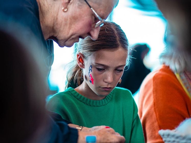 Image of An older woman leans over to help a young girl with face paint on her cheek as they work on a craft activity together at a table, surrounded by other people.