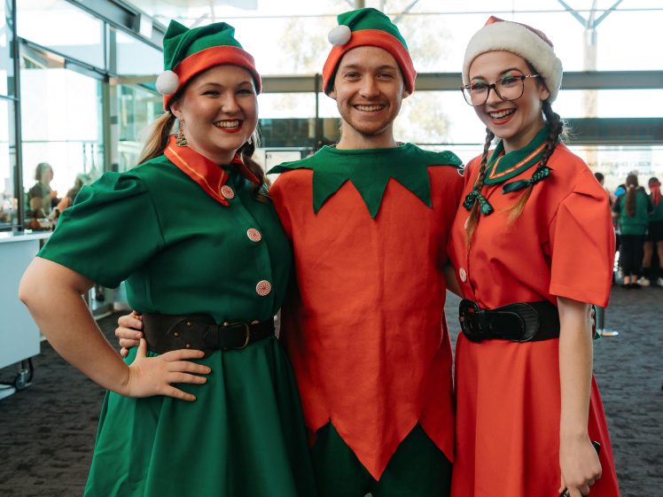 Image of Three people dressed in festive Christmas costumes—two women as elves in green and red dresses, and a man in an orange elf outfit—smile and pose together indoors with holiday decorations visible.