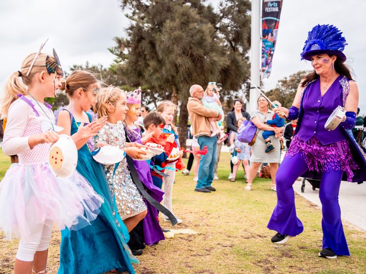 Image of A woman in a bright purple costume and hat leads a group of smiling children dressed in colourful costumes outdoors, as onlookers watch and take photos at a lively festival.