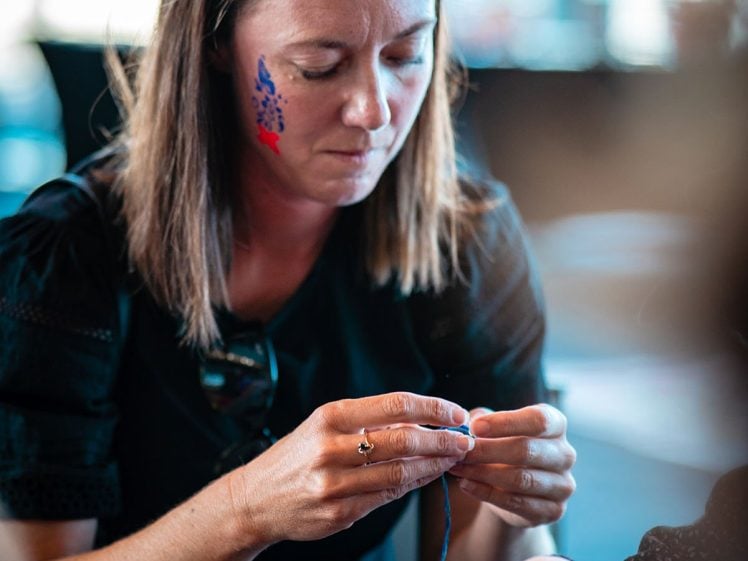 Image of A woman with long brown hair and a colourful painted design on her cheek concentrates on threading beads, while another person’s hands reach into a bowl of beads on the table.
