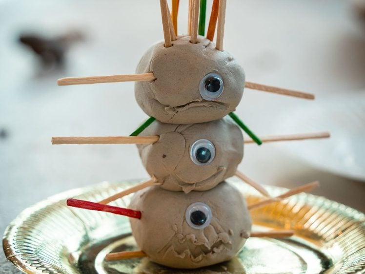 Image of Three clay balls stacked on a gold plate, each with a googly eye and colourful sticks poking out, resembling a playful, abstract creature or sculpture.