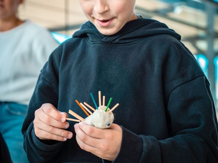Image of A child in a grey beanie and dark hoodie is smiling while making a craft with colourful sticks and a round base. Another person is blurred in the background, and the setting appears to be indoors with large windows.