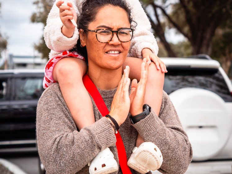 Image of A woman carries a young girl on her shoulders outdoors. The girl wears a colourful paper crown and holds a paper object, while the woman claps her hands. Trees and parked cars are visible in the background.