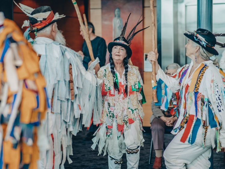 Image of A group of people in colourful, fringed costumes and hats with feathers perform a traditional dance indoors, holding wooden sticks, whilst others watch in the background.