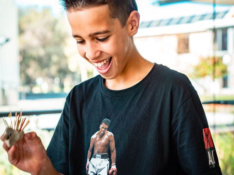 Image of A smiling boy with short dark hair holds a small craft project. He wears a black T-shirt featuring a famous boxing scene. The background shows large windows with sunlight and trees outside.