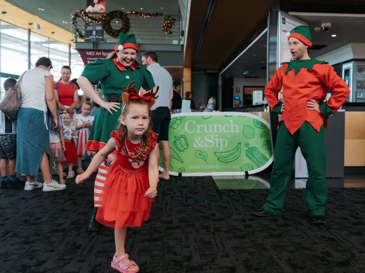 Image of A young girl in a festive dress poses in front of two adults dressed as Christmas elves, whilst other people and children gather in the background near a Crunch & Sip sign.