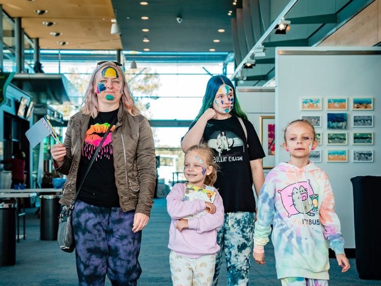 Image of Four people, including two adults and two children, walk indoors wearing colourful, tie-dye and patterned clothes with painted faces and playful accessories. The background shows a bright, modern corridor with large windows.