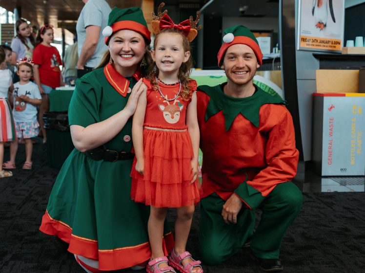 Image of A young girl in a red dress with reindeer antlers stands smiling between two adults dressed in festive elf costumes at an indoor holiday event.
