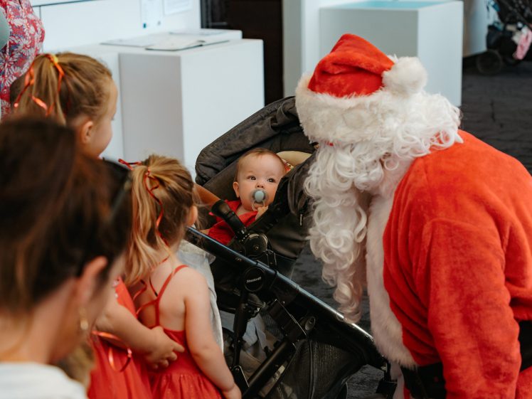 Image of A person dressed as Father Christmas bends down to greet a baby in a pushchair, surrounded by young children in festive red outfits. The baby is looking up at Father Christmas while holding a dummy.
