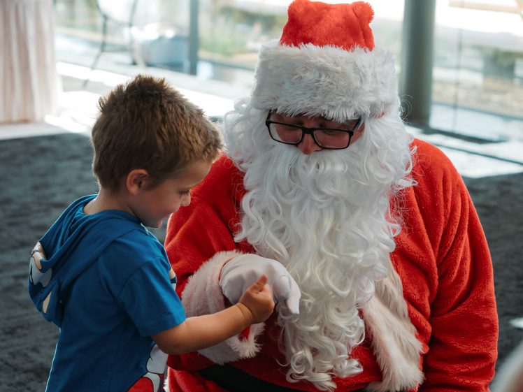 Image of A young boy wearing a blue shirt fist-bumps a person dressed as Father Christmas, who is sitting and smiling. They are indoors with sunlight streaming through large windows in the background.