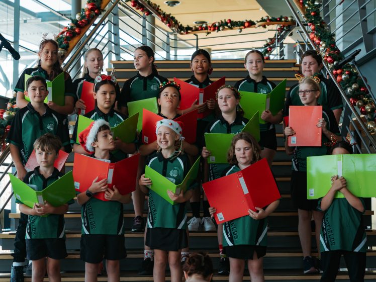 Image of A group of children in green uniforms stand on a decorated staircase, holding colourful songbooks, some wearing festive headbands, singing together in a holiday performance.