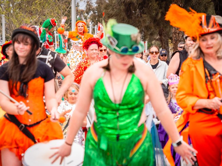 Image of A lively parade with people in colourful costumes, including orange, green, and yellow outfits. Some participants play drums, whilst others walk behind, smiling and enjoying the festive outdoor event.