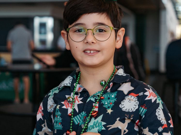 Image of A young boy wearing glasses and a festive shirt stands indoors, smiling. He has a green balloon hat with a cartoon drawing on top and colourful beaded necklaces around his neck.