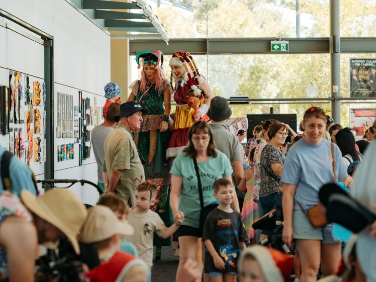 Image of A busy indoor event with adults and children walking through a corridor. Two performers on stilts dressed in colourful costumes stand out amidst the crowd. Art is displayed on the wall to the left.