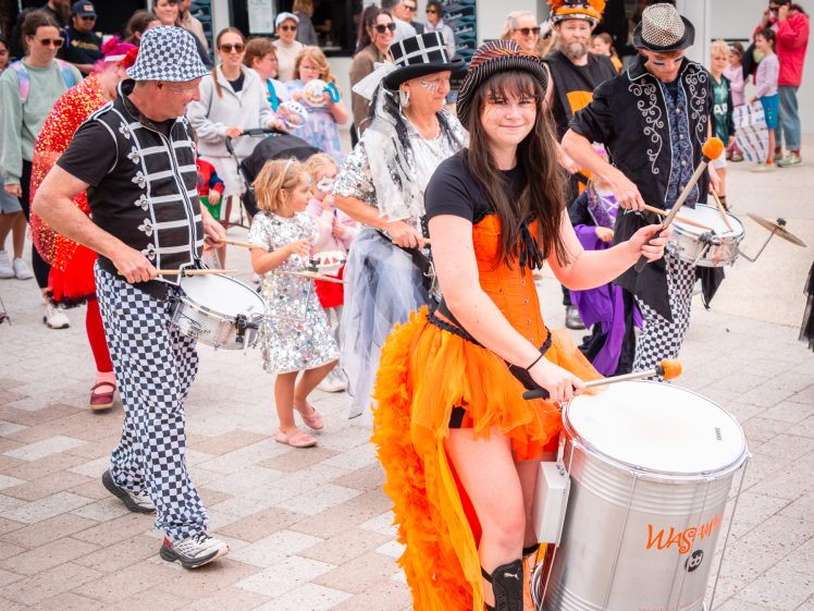 Image of A lively street parade features drummers in colourful costumes, led by a woman in an orange dress. A crowd, including children, follows and watches. The scene is festive, with a takeaway shop in the background.